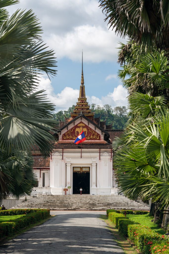 A path leads to the Royal Palace Museum in Luang Prabang, framed by palm trees under a blue sky with scattered clouds.