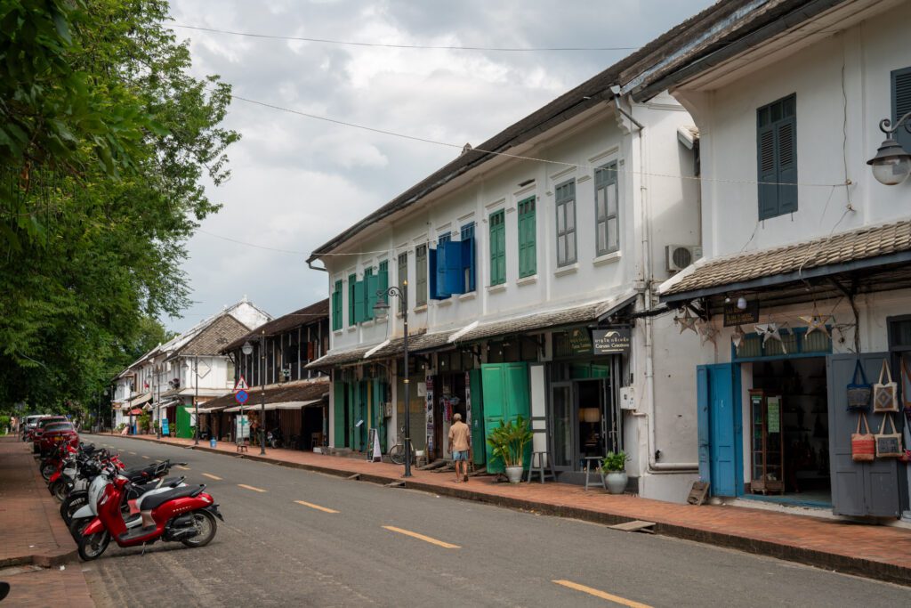 A tree-lined street with colonial-style buildings, scooters parked along the sidewalk, and a person walking. Historic Southeast Asian architecture is prominent.