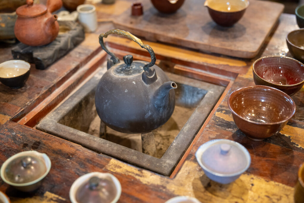 A rustic metal teapot on a sunken stove surrounded by various clay cups and bowls on a wooden table, suggesting traditional tea preparation.