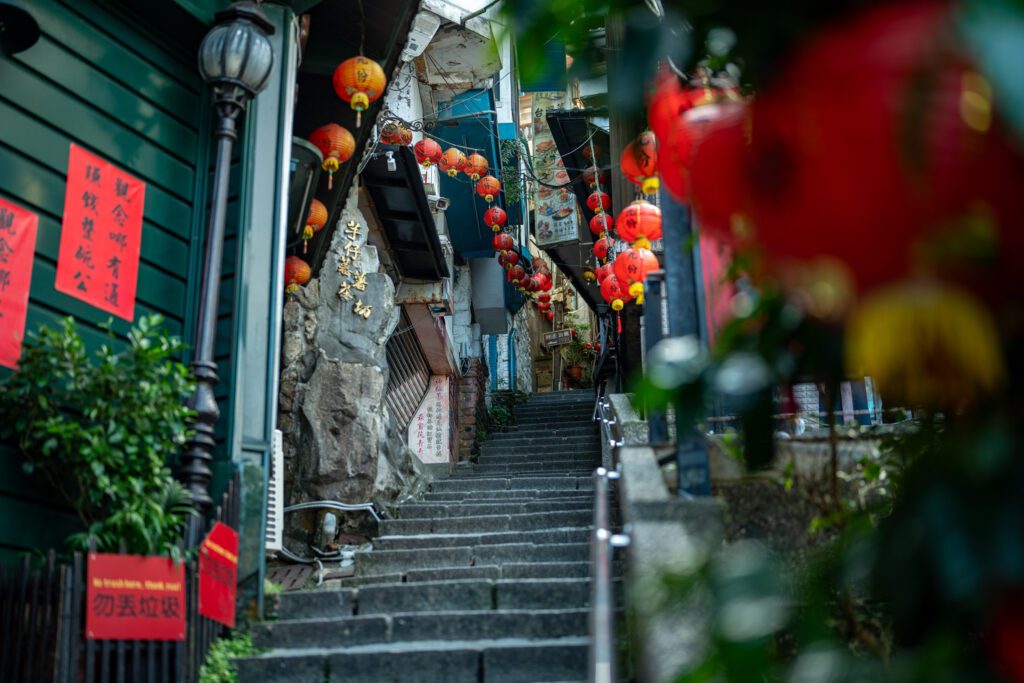 A narrow stone staircase is adorned with red lanterns and vibrant signage, leading up through a bustling alleyway, evoking a traditional Asian atmosphere.