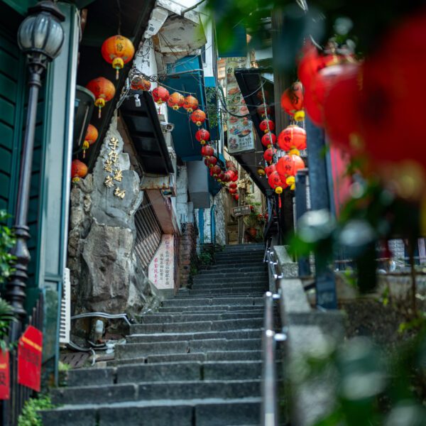 A narrow stone staircase is adorned with red lanterns and vibrant signage, leading up through a bustling alleyway, evoking a traditional Asian atmosphere.