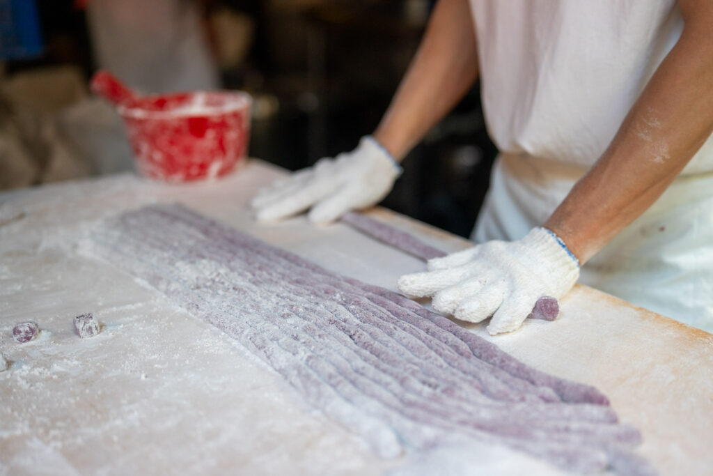 A person wearing gloves prepares long strands of dough dusted with flour on a table, with a red container in the background.