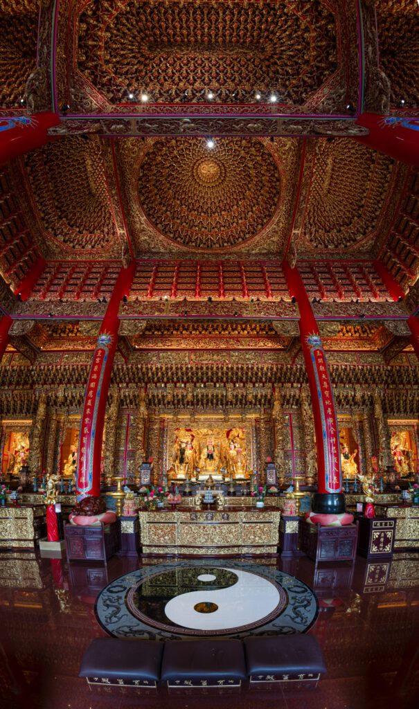 Intricately designed temple interior, featuring ornate gold decorations and statues. Central yin-yang symbol on the floor. Rich red pillars and ceiling detail.