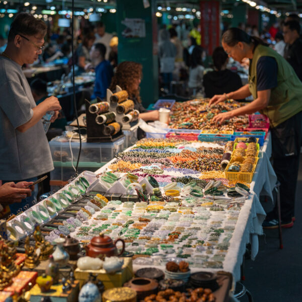 A bustling indoor market with several tables displaying colorful jewelry and trinkets. Two people are browsing and arranging the vibrant items.