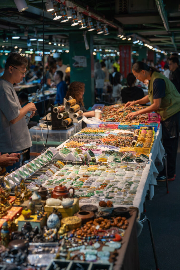 A bustling indoor market with several tables displaying colorful jewelry and trinkets. Two people are browsing and arranging the vibrant items.