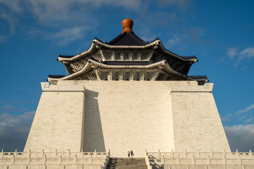 Two people stand before the grand Chiang Kai-shek Memorial Hall, featuring intricate architecture and a clear blue sky background.