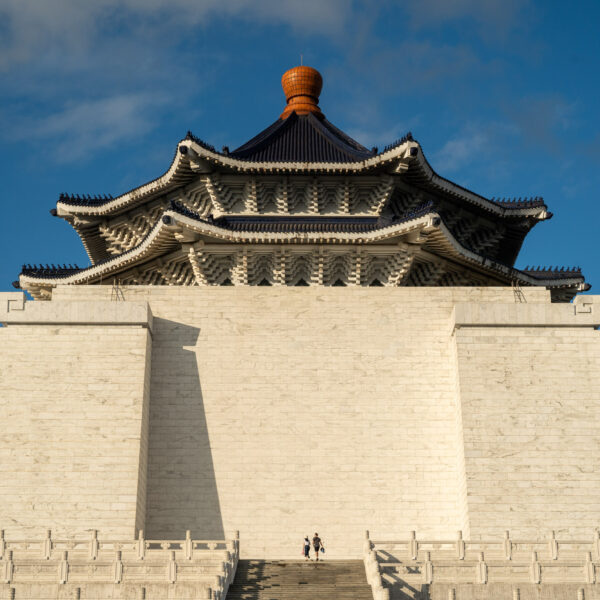 Two people stand before the grand Chiang Kai-shek Memorial Hall, featuring intricate architecture and a clear blue sky background.