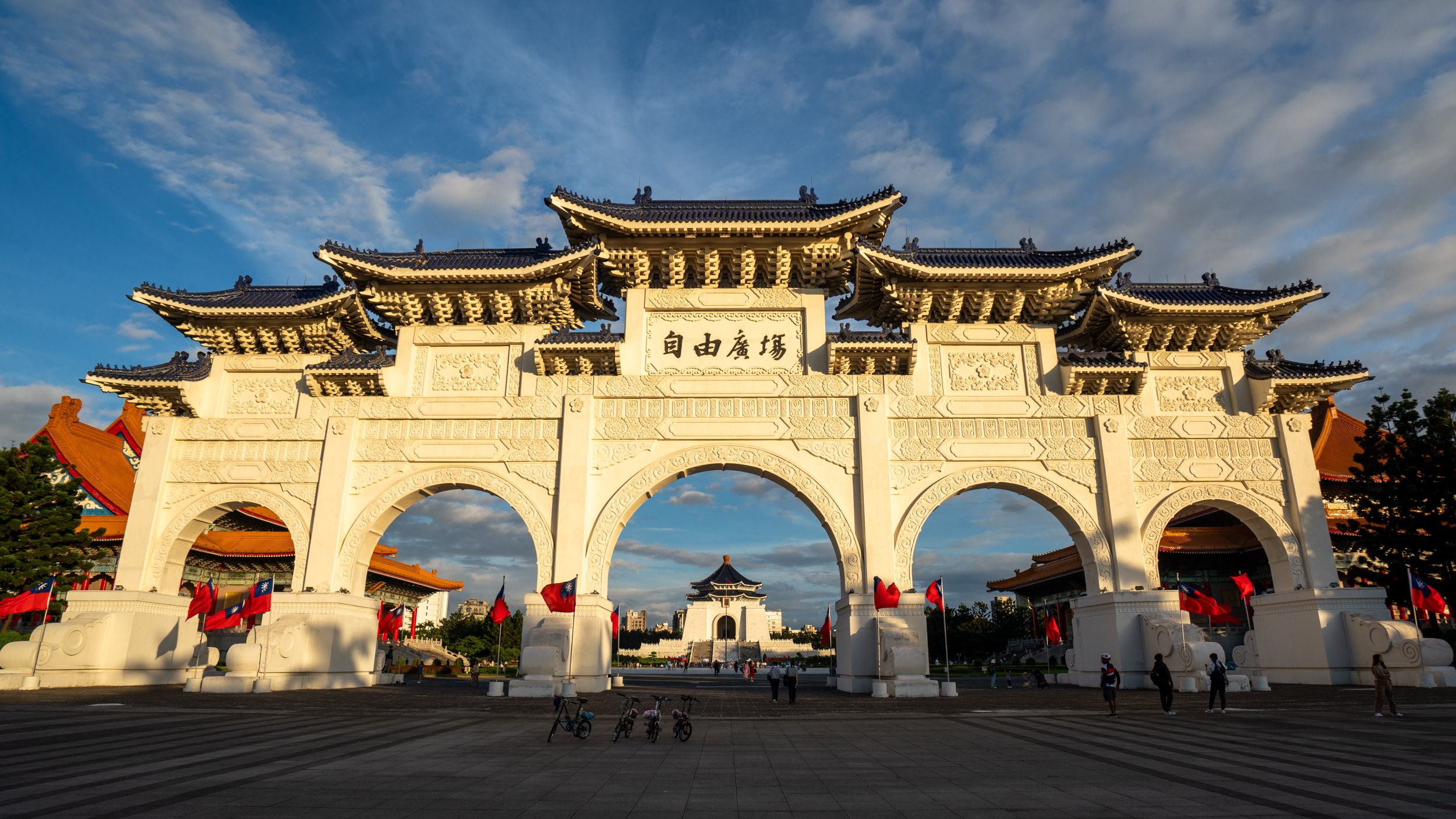 Ornate gateway at Liberty Square in Taipei, bathed in golden light. A few people and red flags are visible around the majestic arches.