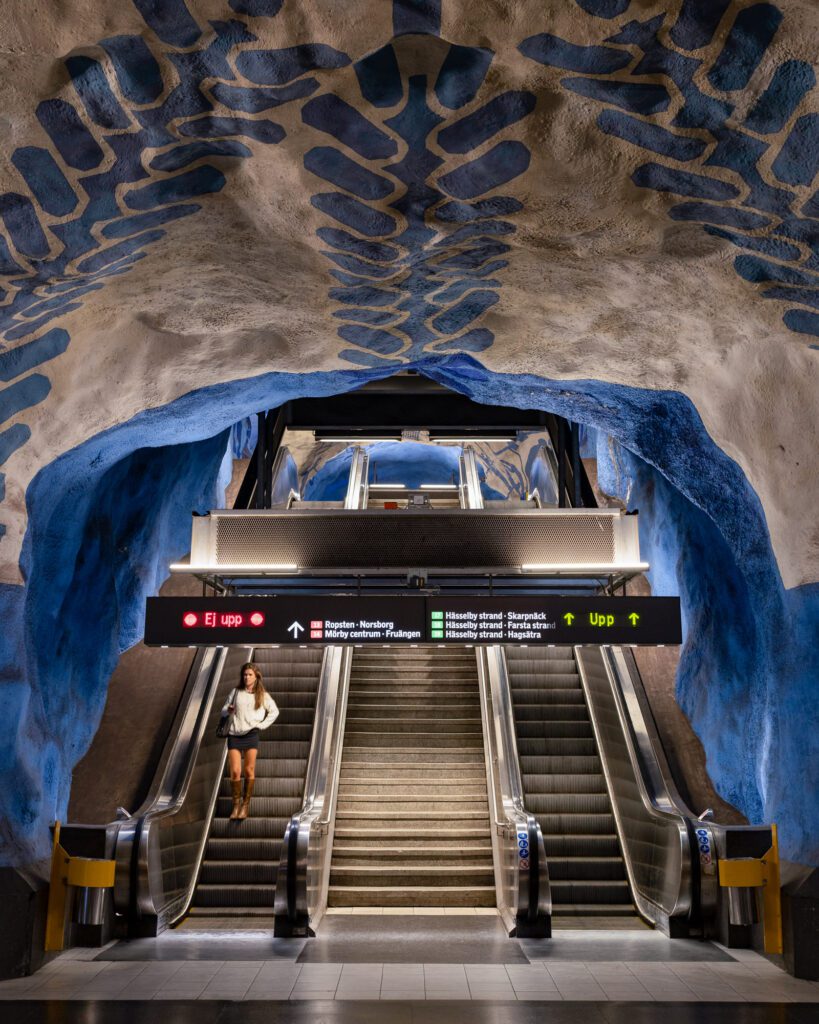 A person stands at Stockholm’s Solna Centrum metro station, featuring striking blue and white artistic cave-like ceiling and an illuminated escalator.