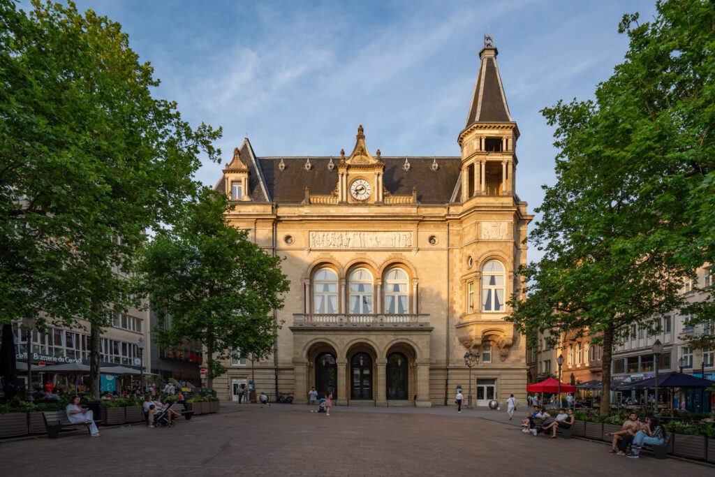 A historic square with people sitting on benches, featuring Cercle Municipal in Luxembourg City, surrounded by trees and vibrant urban life.