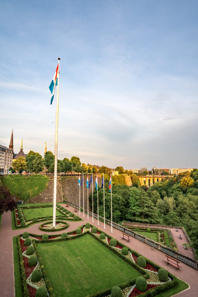 Flags stand in a manicured garden in Luxembourg City, with the Adolphe Bridge and cityscape visible in the background under a clear sky.