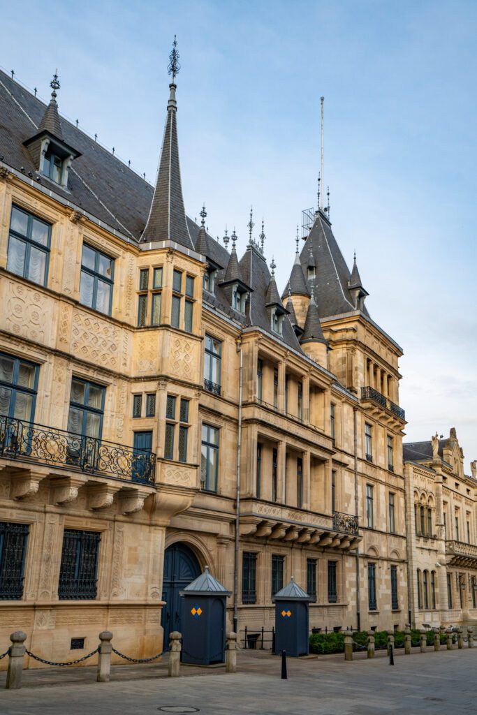 Historic building in Luxembourg City with ornate architecture, gabled roofs, and a stone facade. Two guard posts are visible in the foreground.