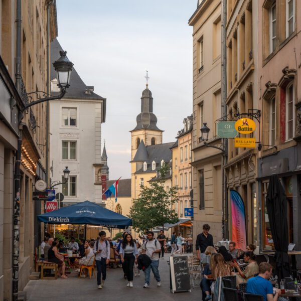 A bustling street with outdoor cafes and people. The spire of Saint Michael's Church in Luxembourg City rises in the background, under a clear sky.