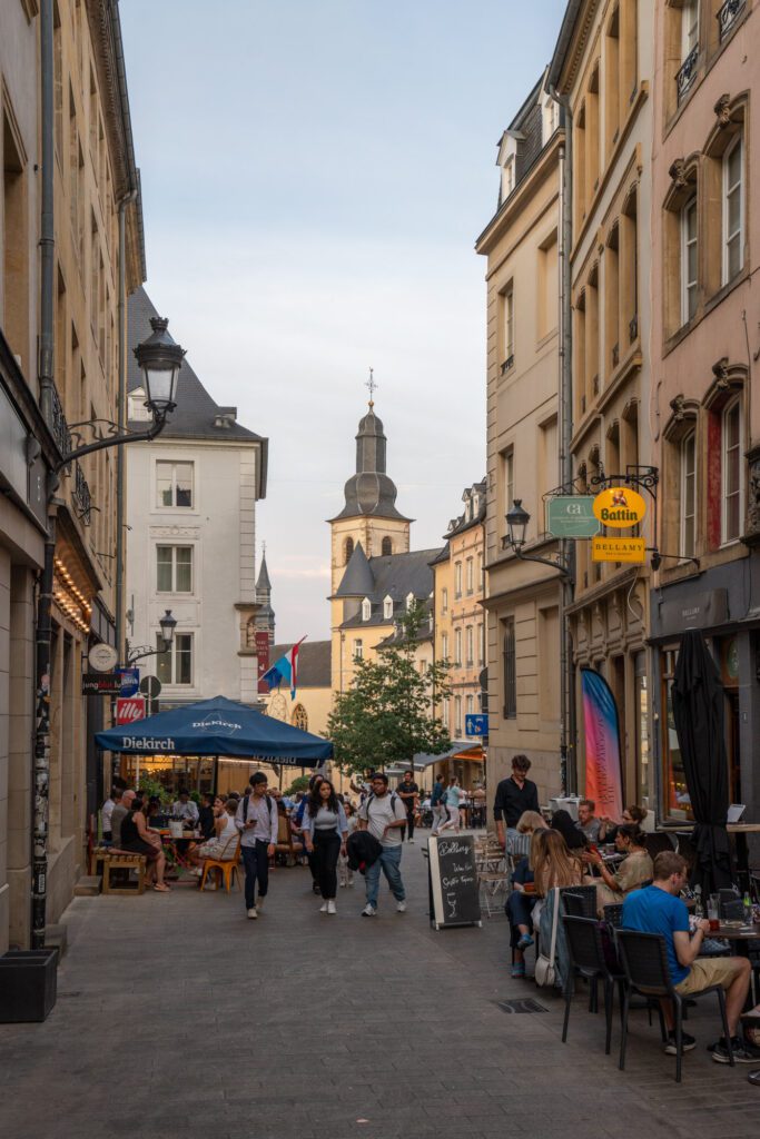 A bustling street with outdoor cafes and people. The spire of Saint Michael's Church in Luxembourg City rises in the background, under a clear sky.