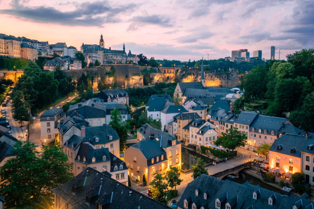 Evening view of Luxembourg City, highlighting historic fortifications and a blend of medieval and modern architecture under a colorful sky. Lush greenery surrounds.