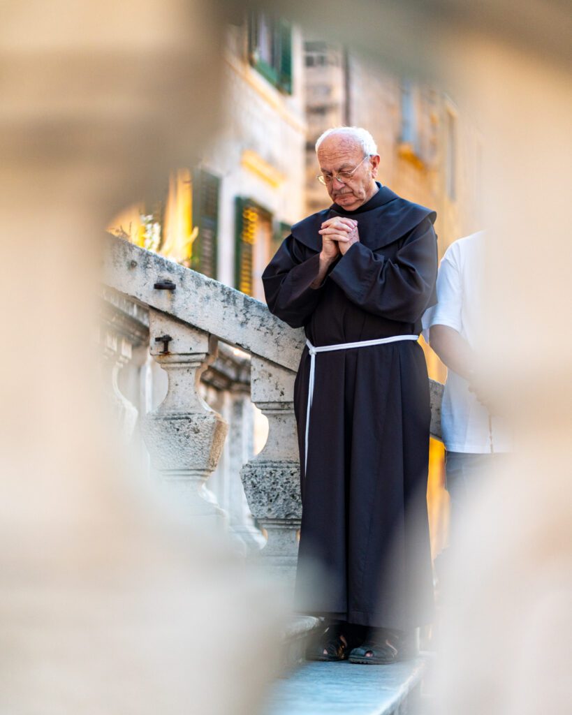 A person in a religious robe stands solemnly on a stone staircase, framed by blurred foreground elements, in a historic European city setting.