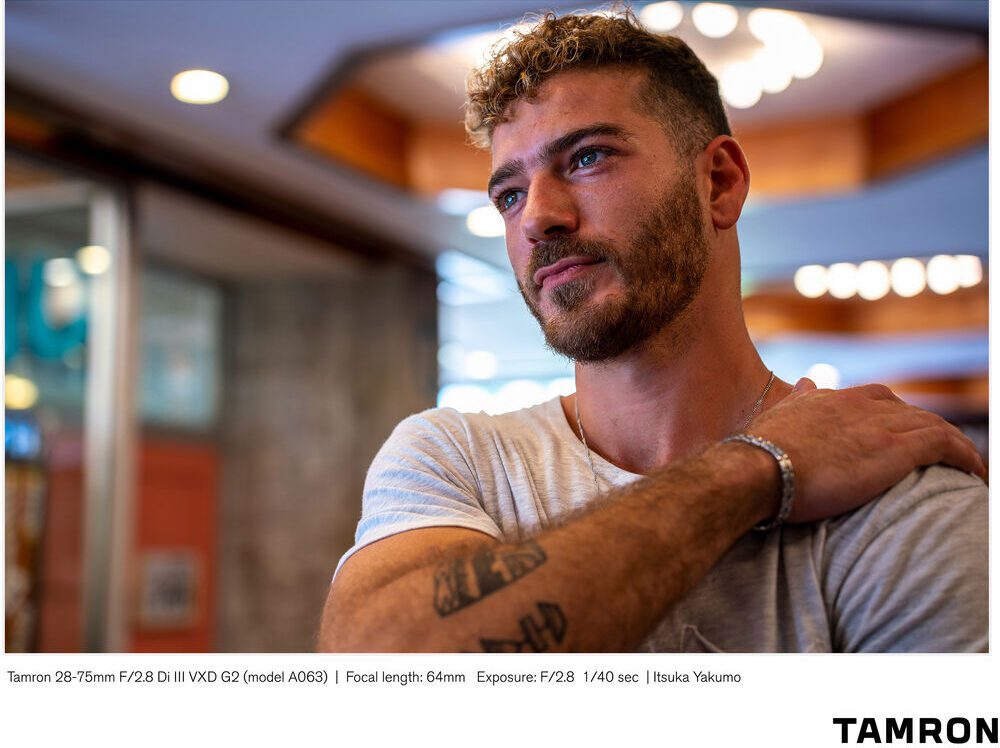 A person with tattoos and a beard poses inside a building with warm lighting, contemplating. Visible text refers to camera details and lens brand Tamron.