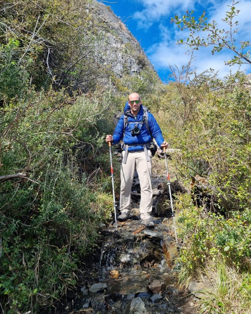 A person hiking with poles on a rocky path surrounded by green foliage and a steep hill under a clear blue sky.