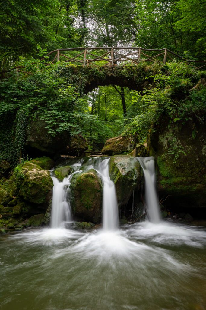 A serene waterfall cascades under a rustic wooden bridge in a lush forest, surrounded by vibrant greenery and moss-covered rocks.