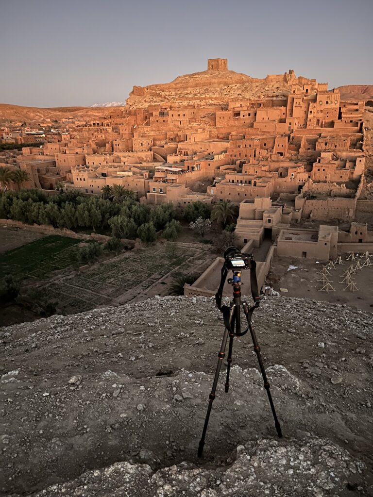 A camera on a tripod overlooks an ancient clay cityscape lit by the setting sun, surrounded by desert landscape. Products for travel photographers