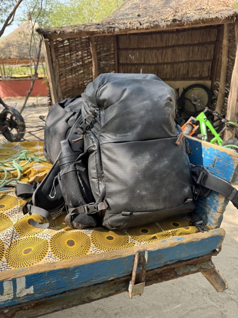 A black backpack on a decorated cart, near a traditional thatched hut; a bicycle is visible in the background.