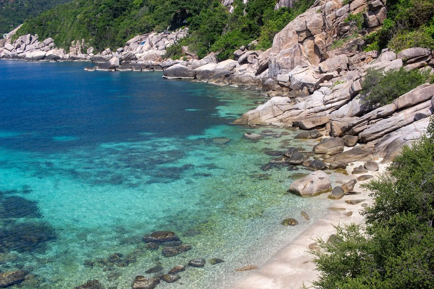 Rocky shoreline with clear turquoise water and lush green landscape in a serene beach setting.